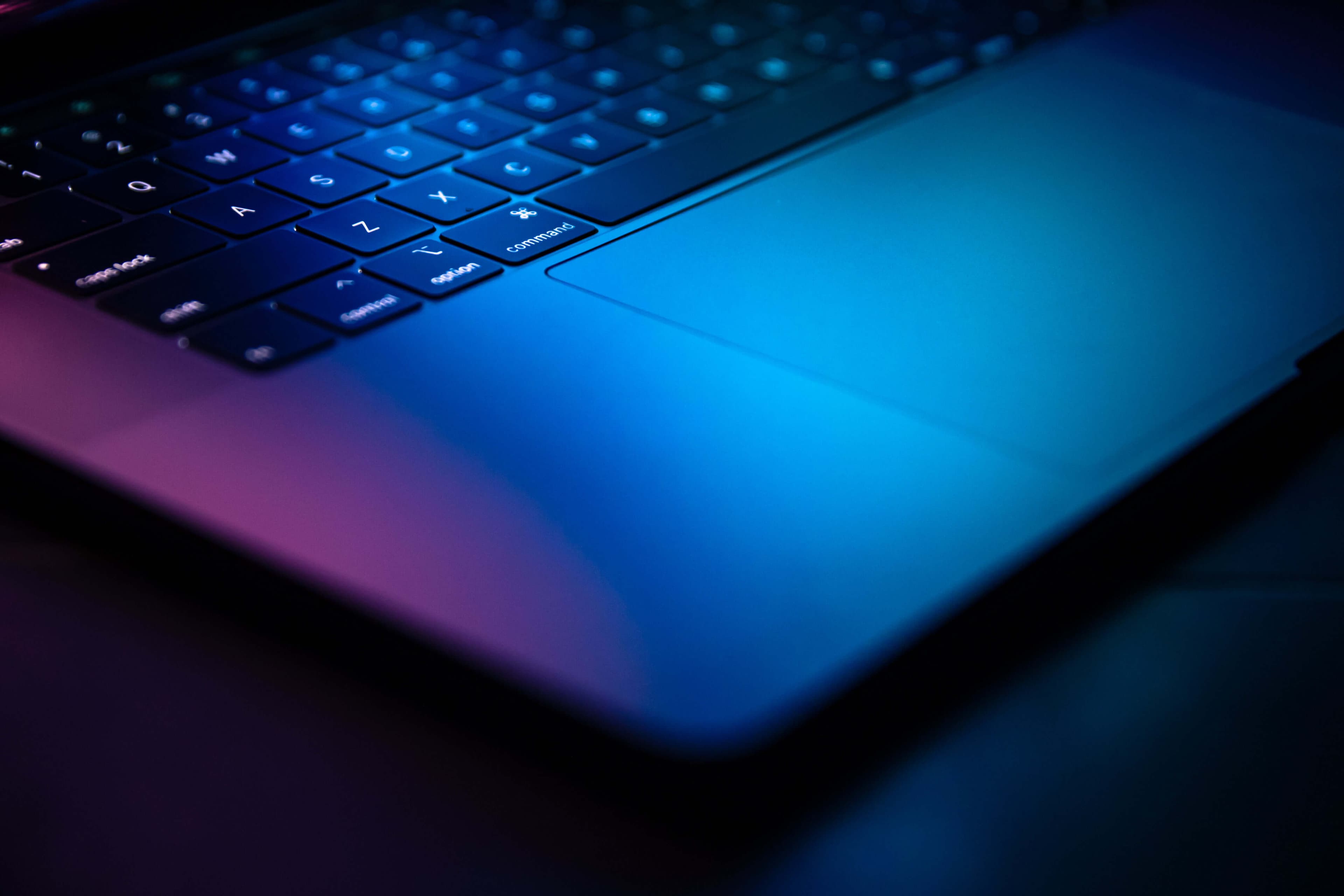 Grey Mac Laptop with a black keyboard with a rainbow gradient light shining down on it from above.