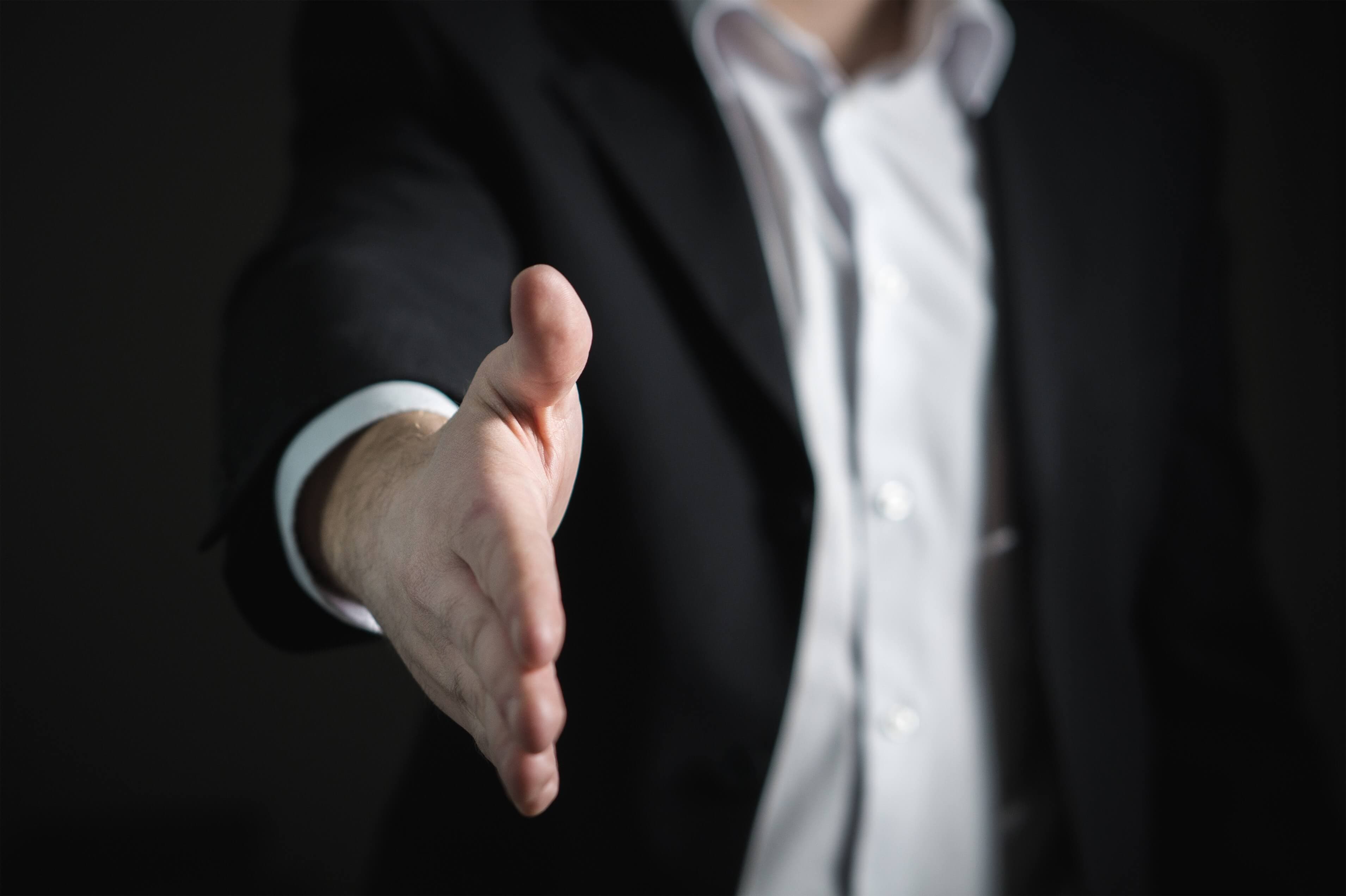 Man in a black suit suit and a light grey shirt that is reaching out his hand for a greeting handshake.