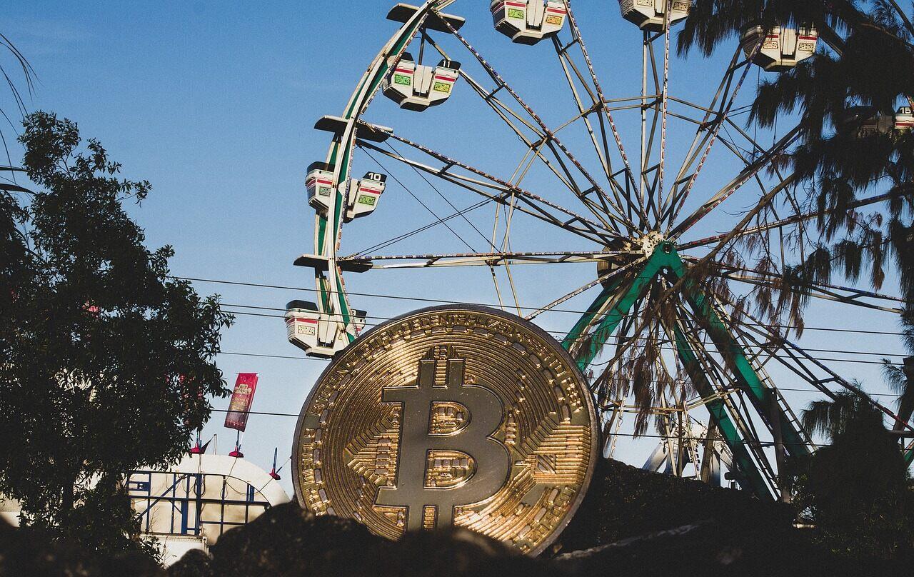 A golden coin with a Bitcoin symbol on it sitting in a pile of dirt with a ferris wheel and an amusement park in the background.