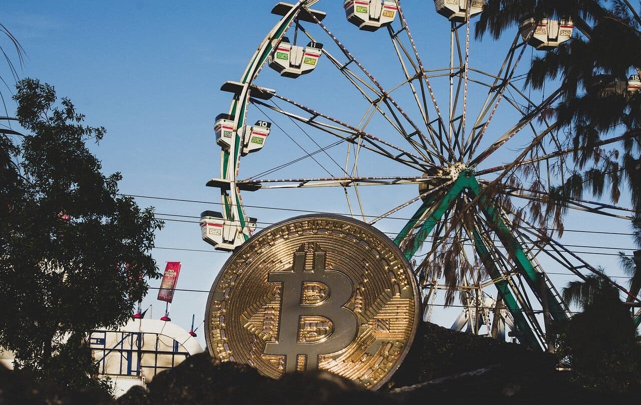 A golden coin with a Bitcoin symbol on it sitting in a pile of dirt with a ferris wheel and an amusement park in the background.