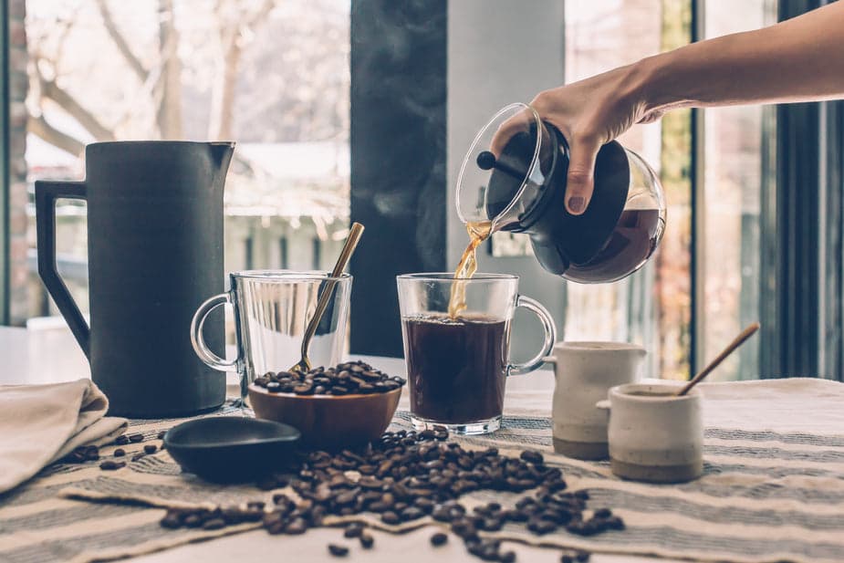 A bunch of coffee beans on a table with someone pouring coffee into a cup from a glass slow drip coffee maker with a black lid.