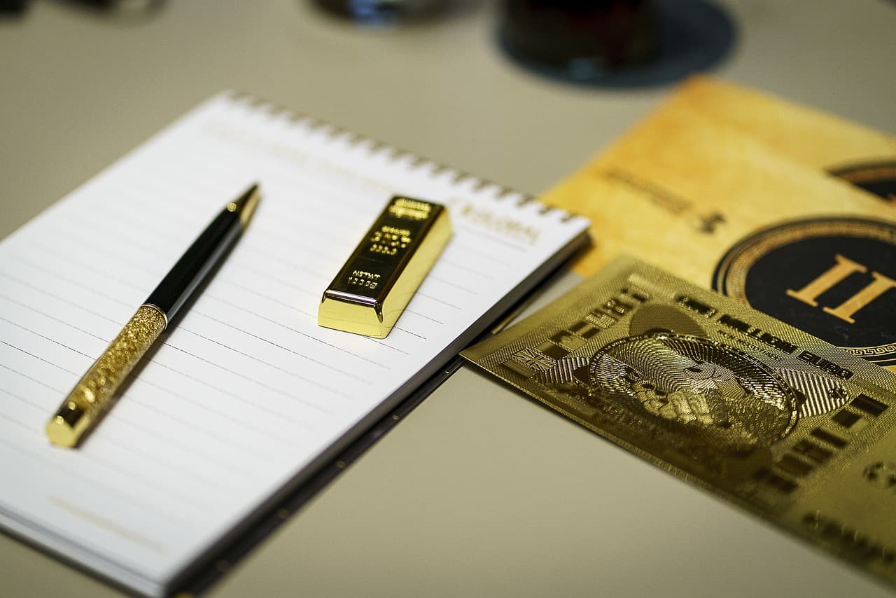 A white lined notebook sitting on a lightly colored table with a golden pen and a golden bar on top of it next to each other.