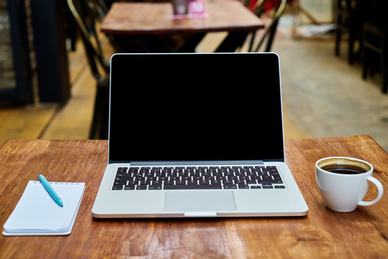 An Apple Macbook sitting on a wooden table turned off next to a cup of coffee and a small notebook with a pen resting on top.