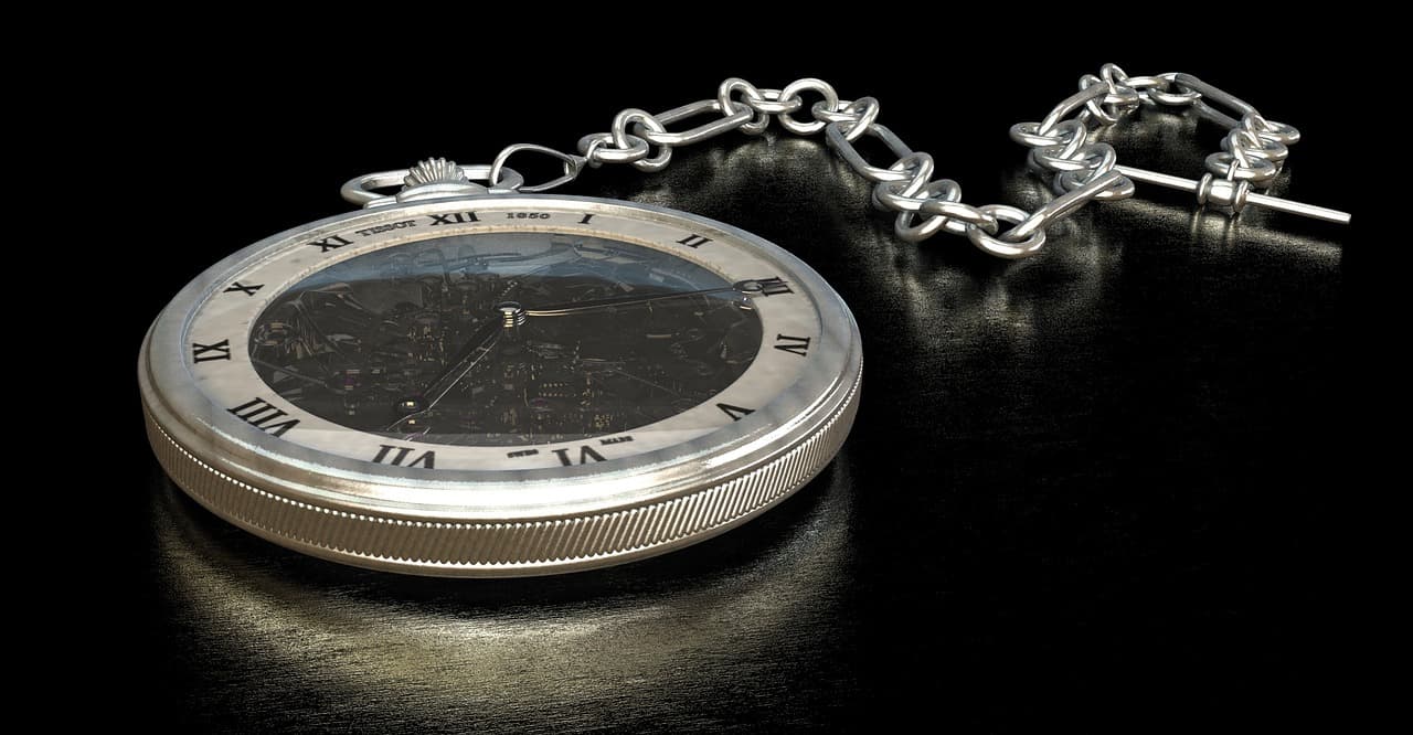 A silver pocket watch and silver chain with a darkly colored dial that has exposed black gears and black hands on a black table.