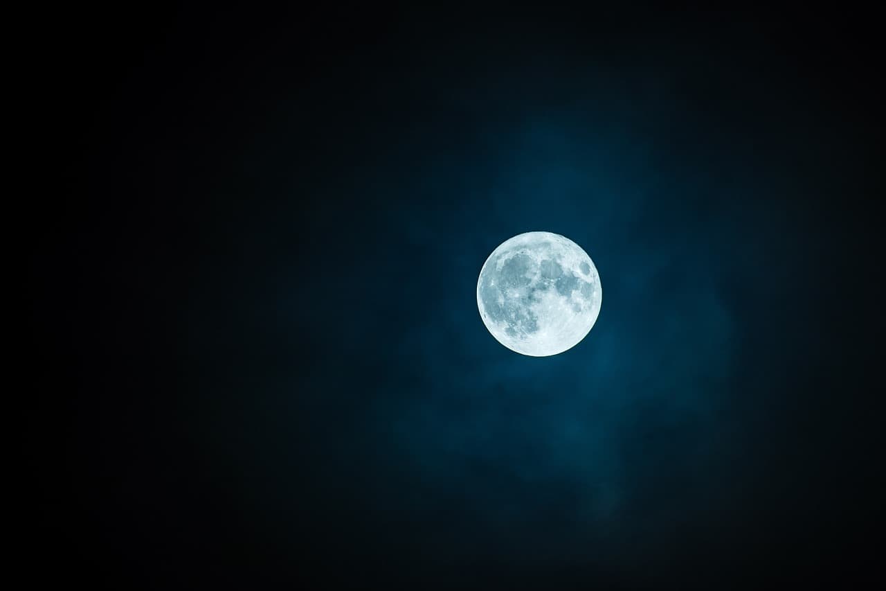 A bright full moon in the night sky is lit up blue and surrounded by blue clouds covered by darkness and other gray clouds.