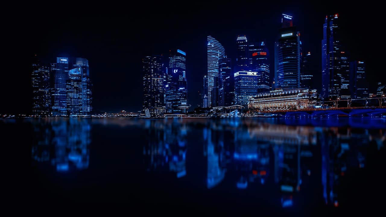 A city on the water is lit up with blue lights and each building has its own white and yellow room lights on in random windows.