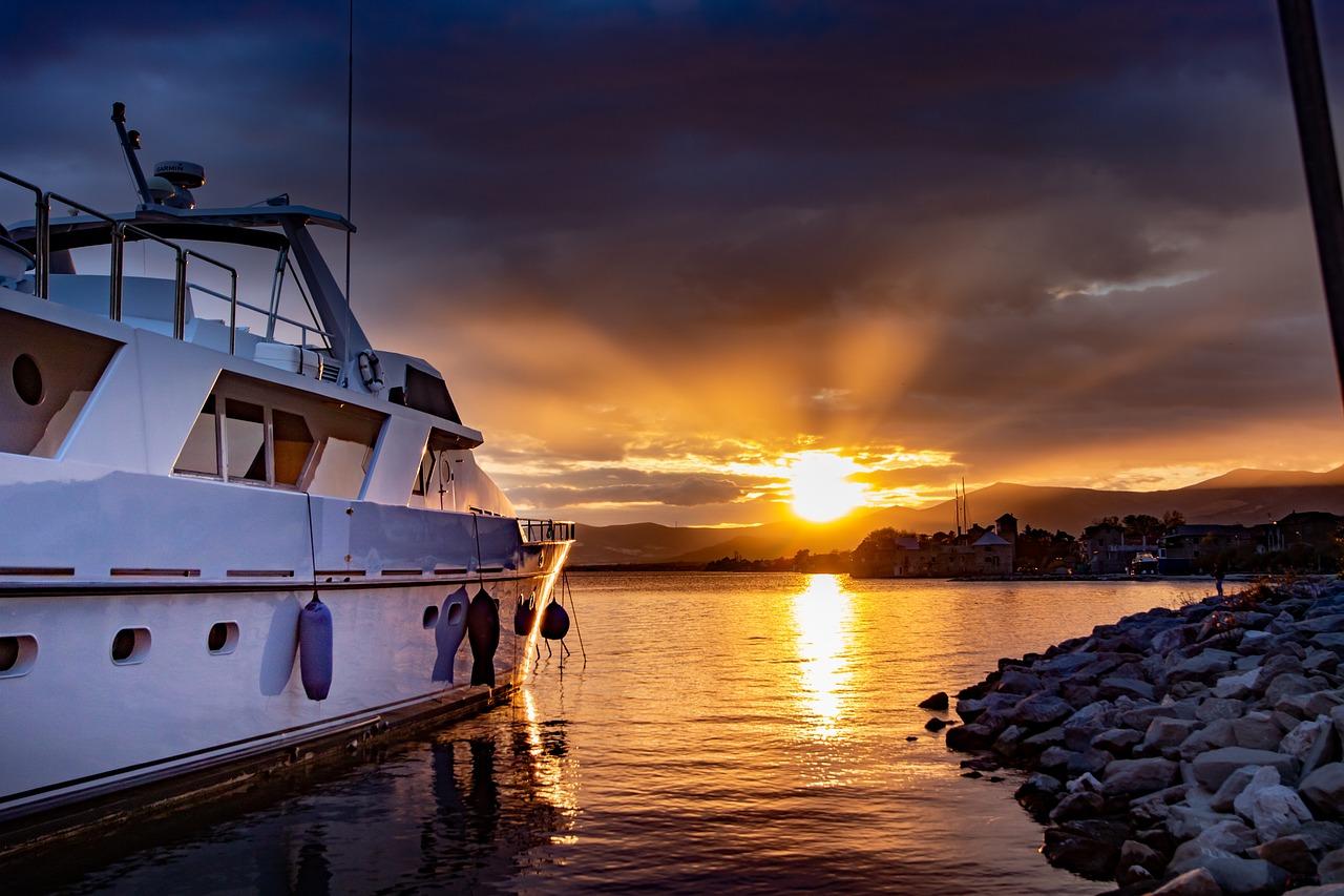 An image of a white yacht in the blue water while the sun sets in the background shining the land with orange and yellow.