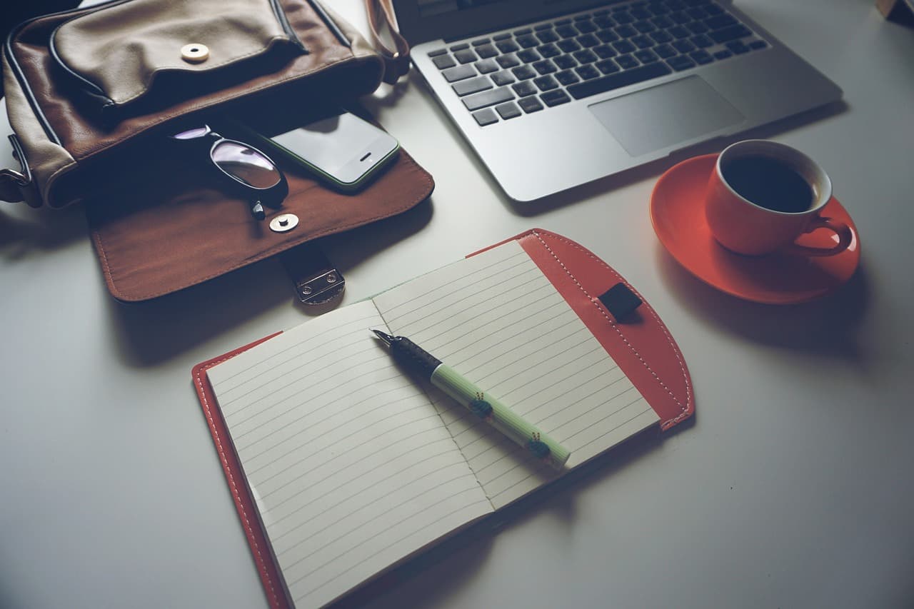 A notebook with a pen on top of it next to and open leather bag, a macbook laptop, and a red coffee cup and matching plate.