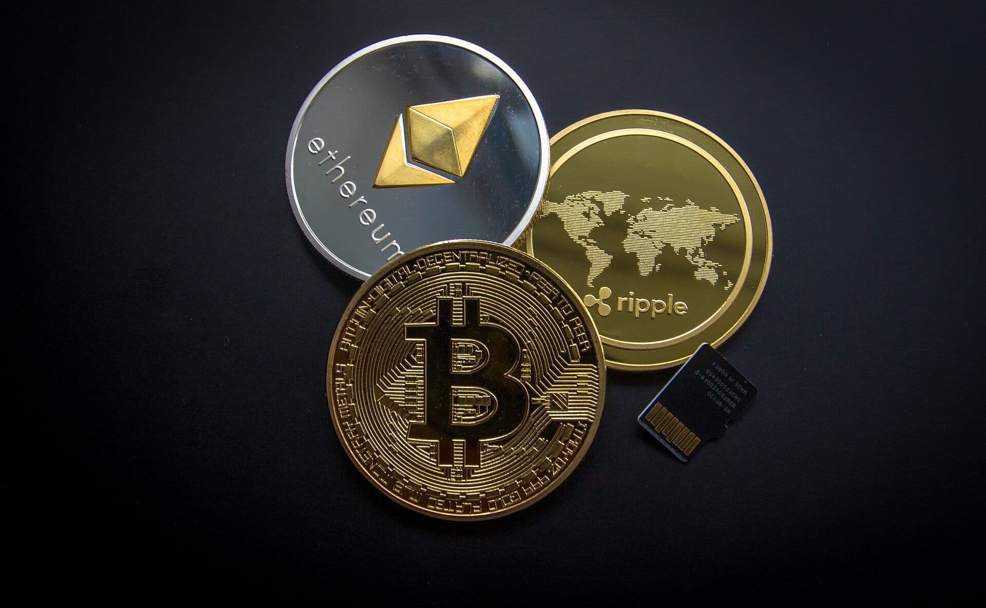 Shiny gold and silver coins of Bitcoin, Ethereum, and Ripple sitting on a black table with a black sd card sitting next to it.