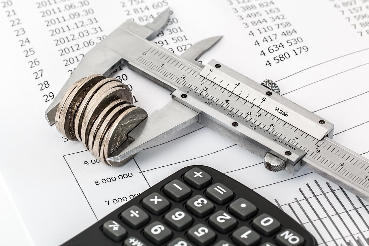 A metal caliper that is holding ten coins of different sizes next to a calculator on a piece of paper that has numbers on it.