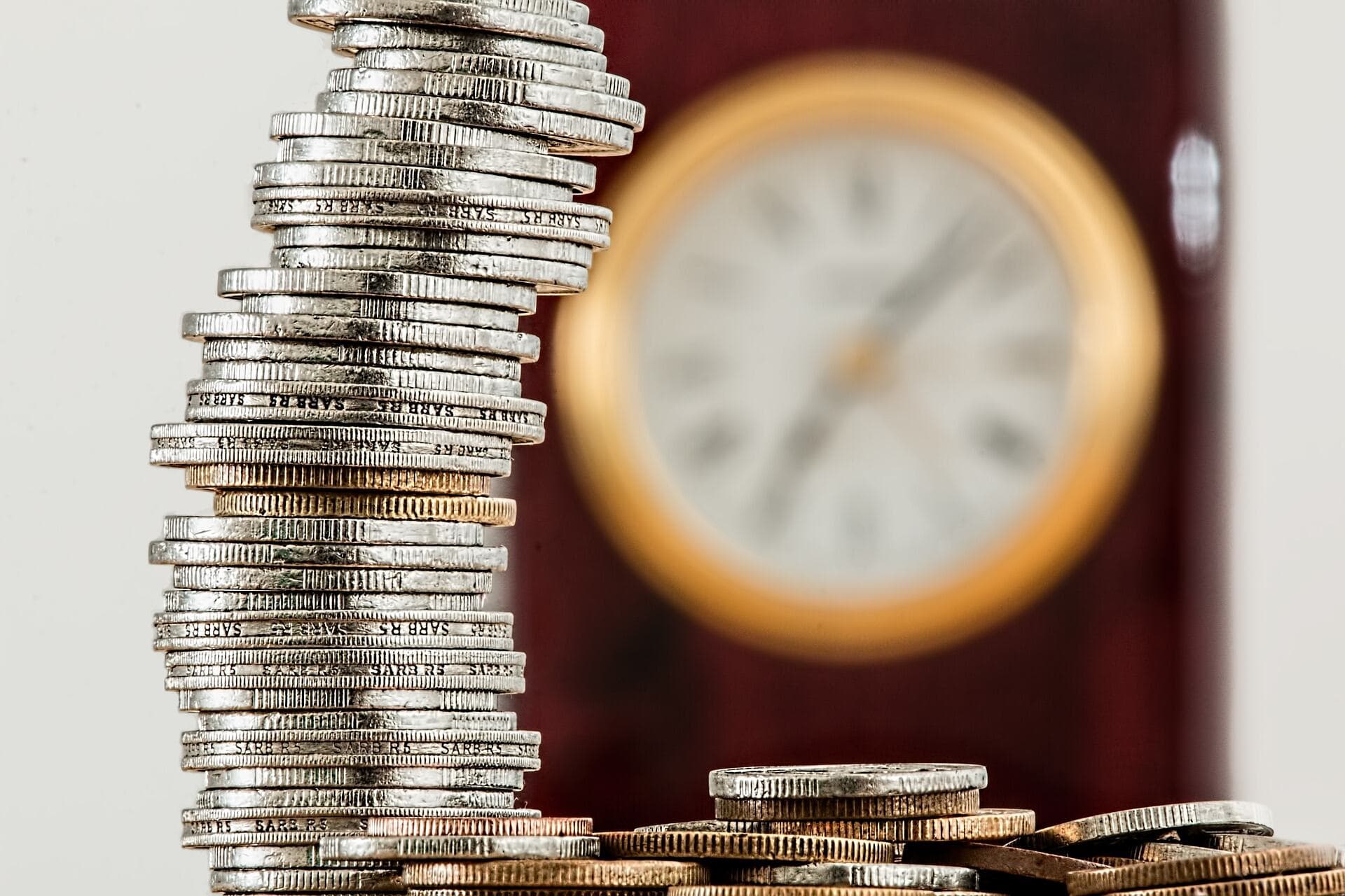 A single stack of silver coins that is sitting on top of more unorganized coins that are on a table with a clock behind them.