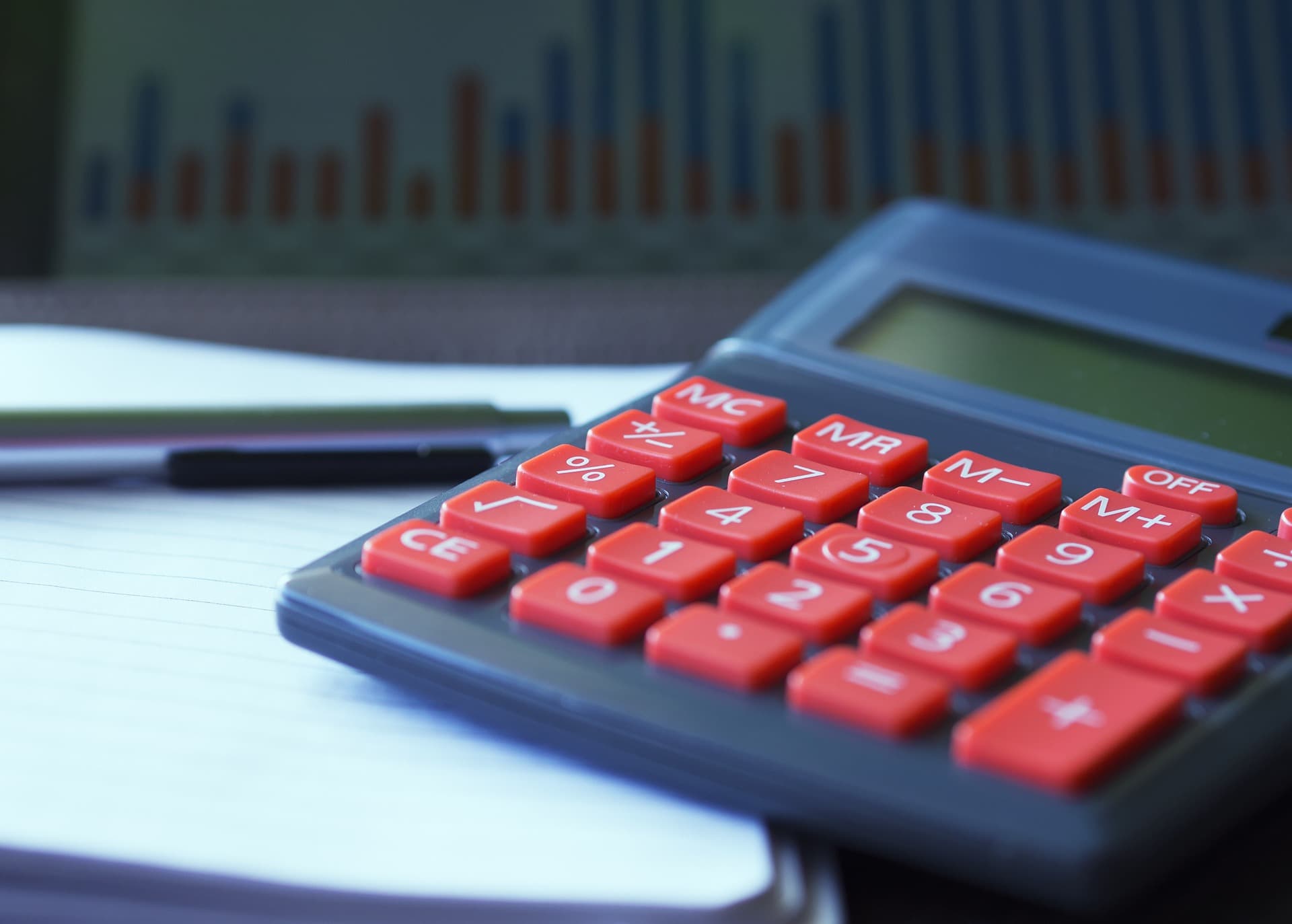 A black calculator with red buttons that is resting on top of a lined notebook that has a pen on top and a chart behind them.