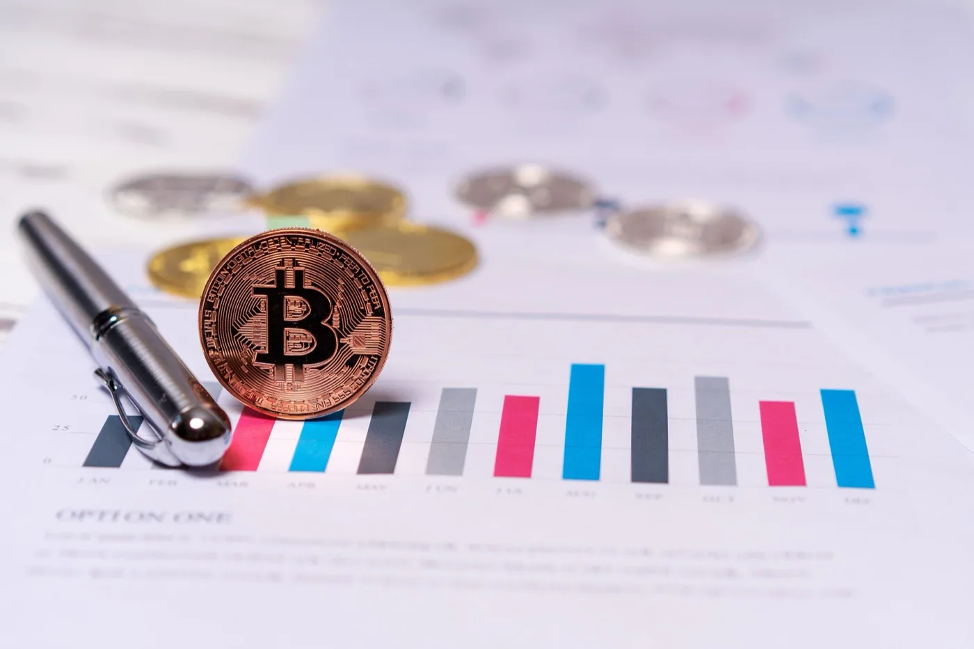 A golden Bitcoin crypto coin standing up on it’s side next to a shiny silver pen with a cap on it all on top of a piece of paper.
