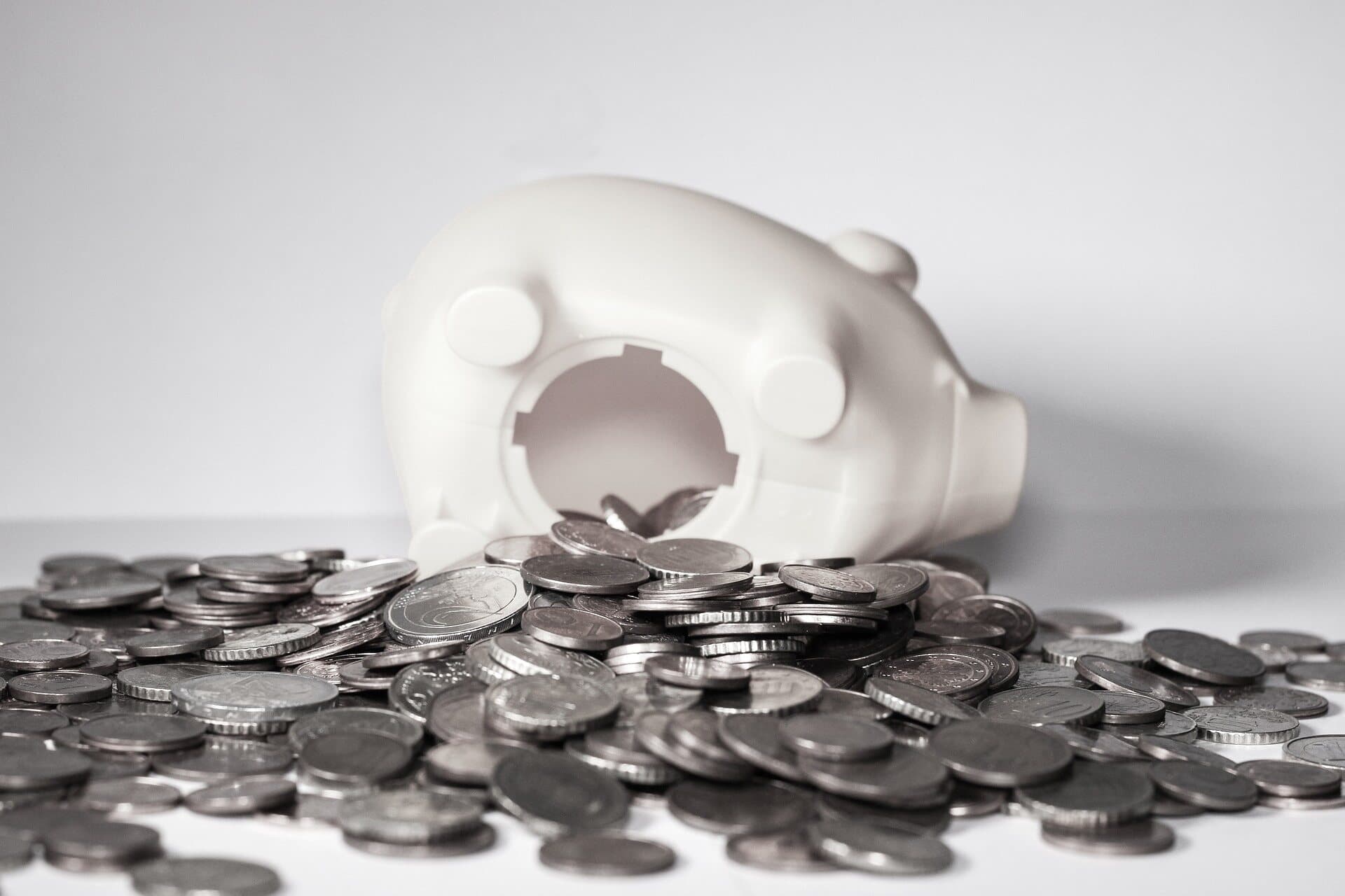 A white plastic piggy bank on its side with the hole on the bottom undone with a large pile of silver coins surrounding it.