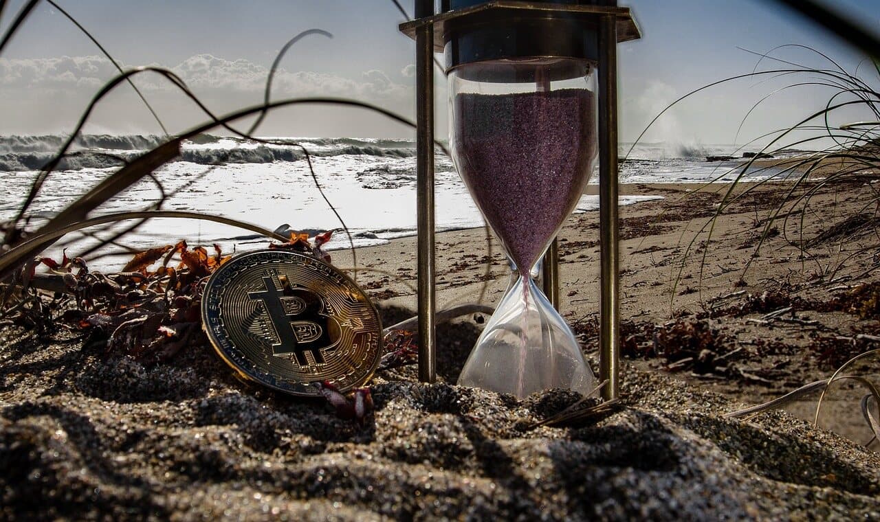 A golden Bitcoin crypto coin in the sand by the beach with an hourglass sitting next to it along with some sticks and leaves.