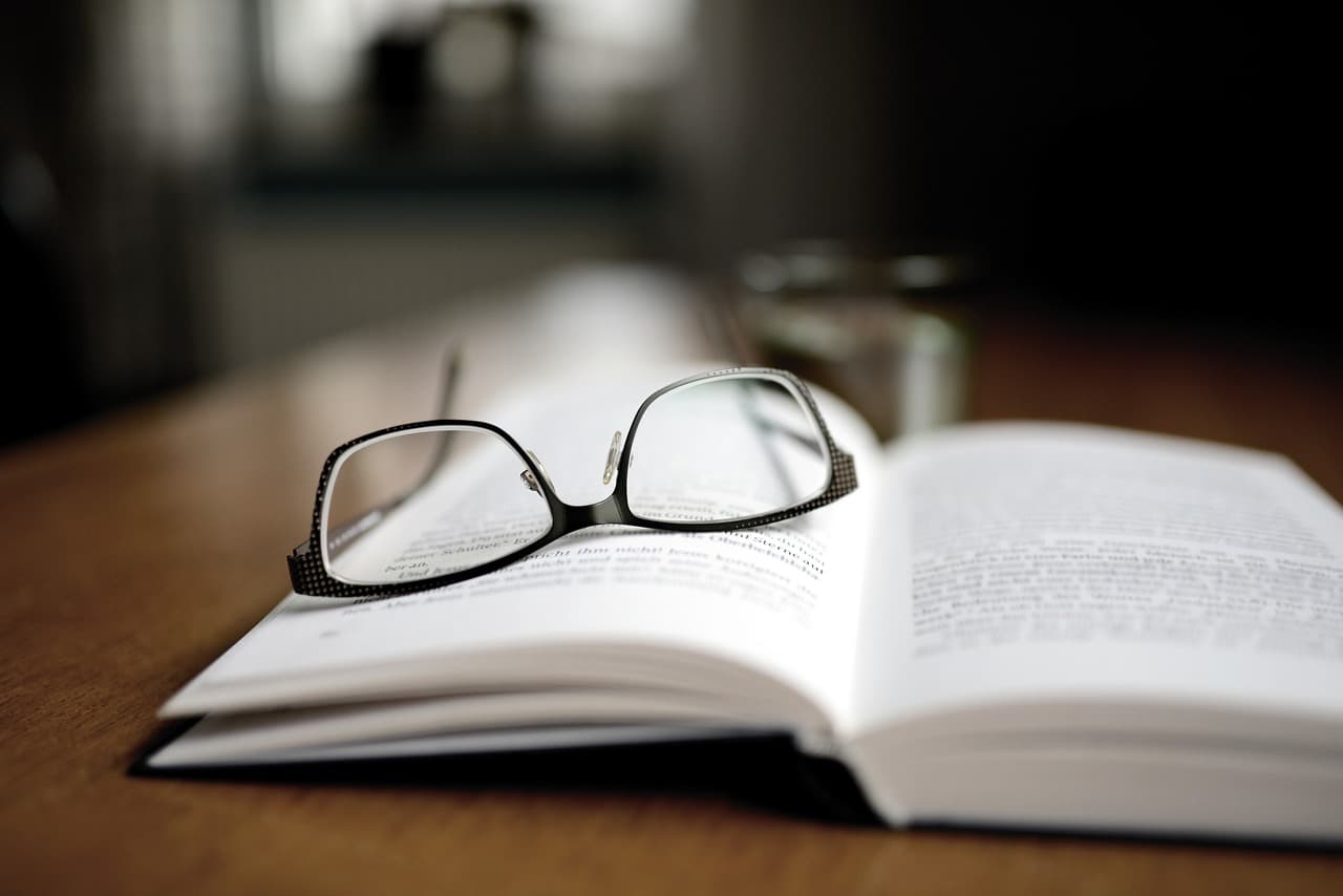 A pair of black glasses on the pages of an open book with a black cover that are all on a wooden table that is a little blurry.