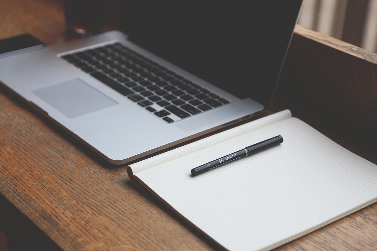 A silver Apple Macbook on a wooden desk next to a notebook that is folded open with a black pen resting on one of the pages.