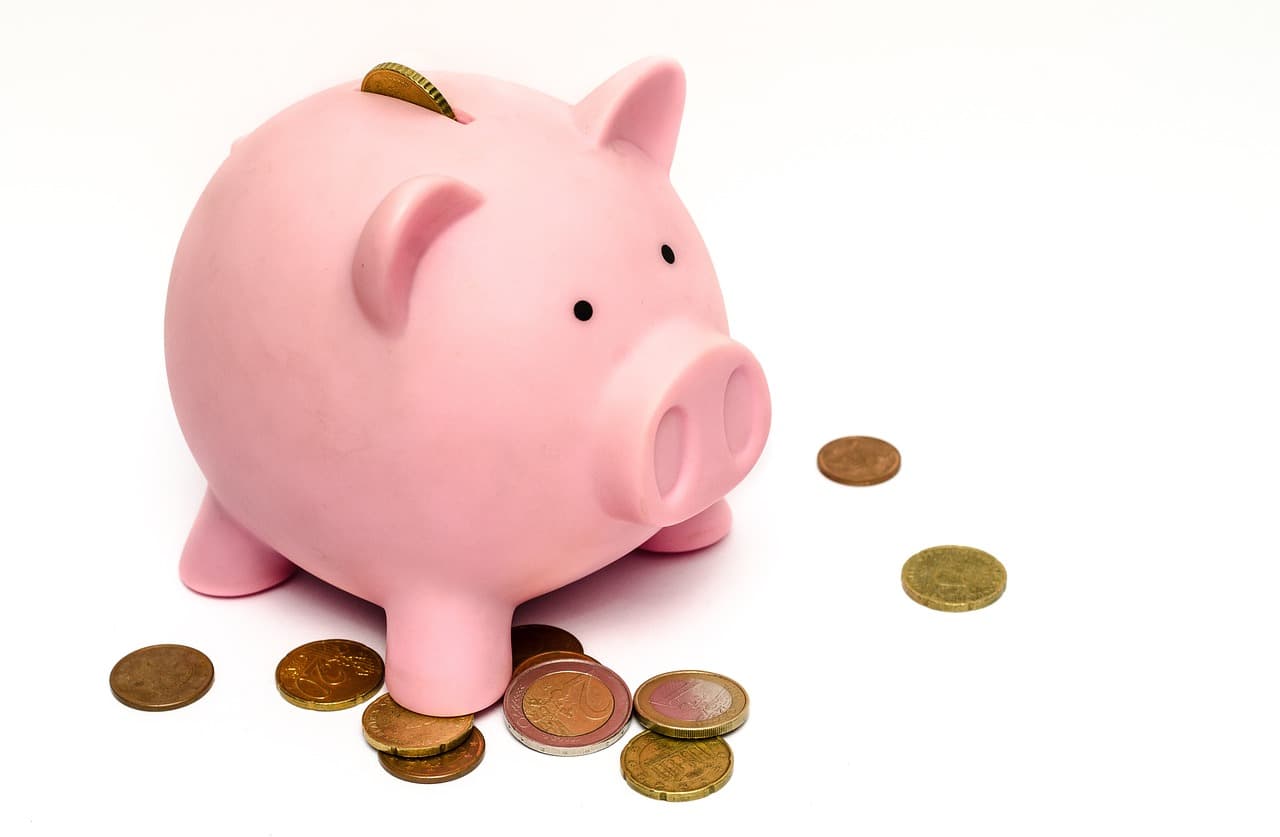 Pink piggy bank that has a coin inside of its slot surrounded by ten other bronze colored coins while on two of them on a table.