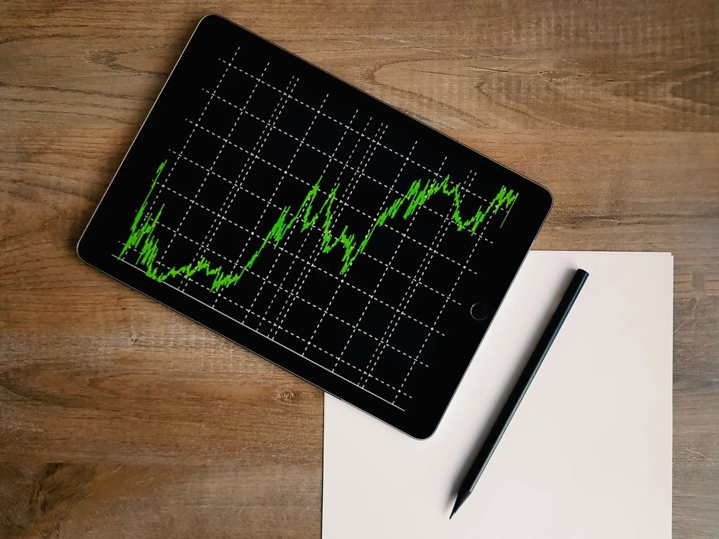 The stock market graph on the screen of an IPad in the color green with a pen and piece of paper sitting next to it.