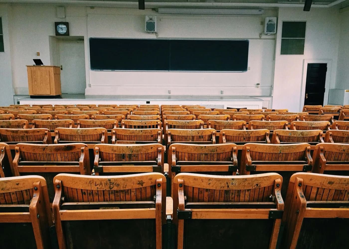 Empty classroom with wooden chairs and a large chalkboard at the front with a podium on the left side of it.