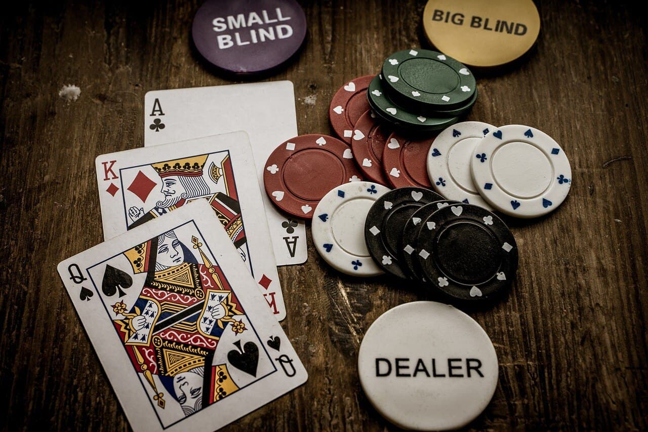 Three cards being a king, queen, and ace next to multicolored poker chips in a pile on top of a darkly colored wooden table.