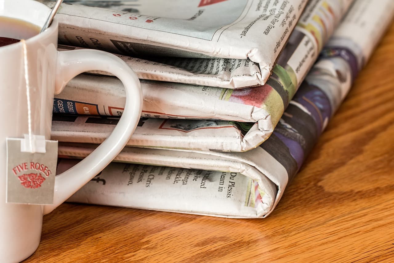 Three stacks of folded newspapers stacked on top of each other next to a coffee cup on top of a lightly colored wooden table.