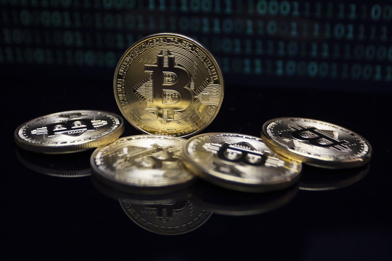 A golden Bitcoin crypto coin sitting on its side surrounded by four other coins on a black table with ones and zeros behind it.