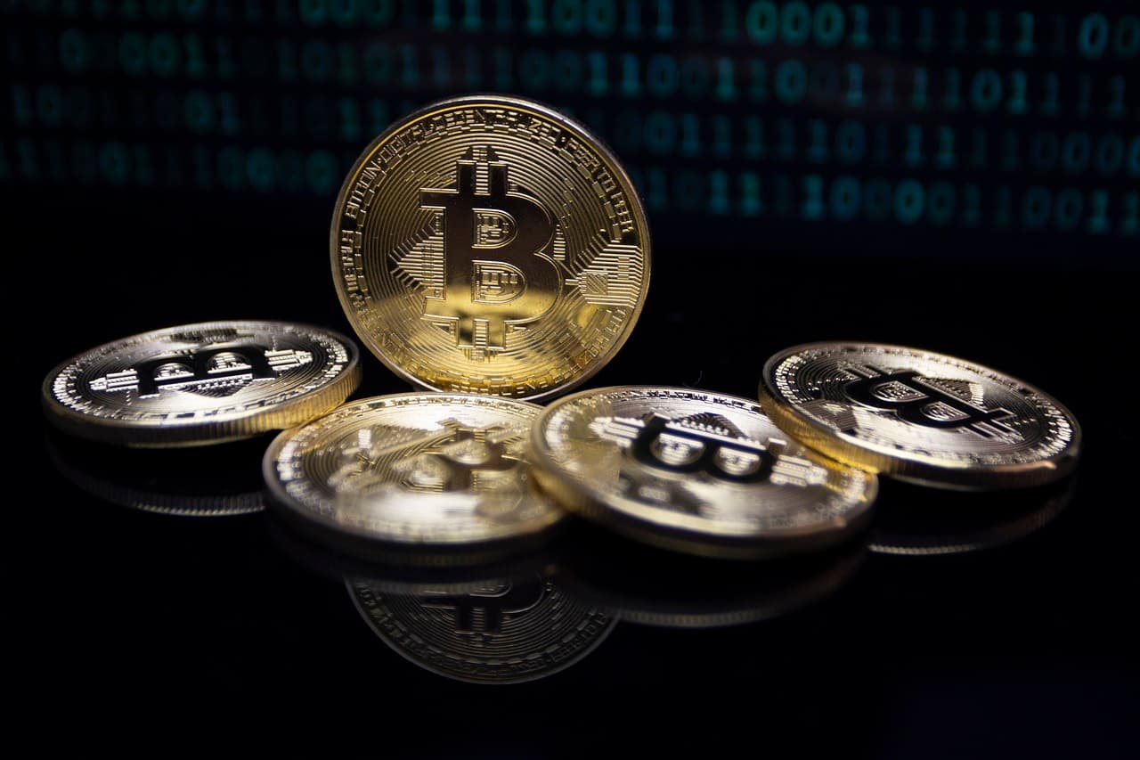 A golden Bitcoin crypto coin sitting on its side surrounded by four other coins on a black table with ones and zeros behind it.