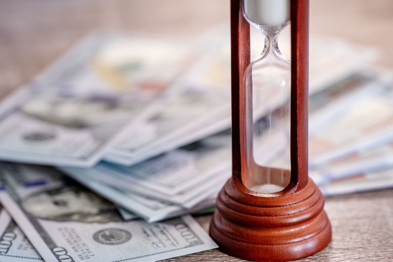 A wooden hourglass with all the sand on the top next to a stack of one hundred dollar bills placed in a fan shape on a table.