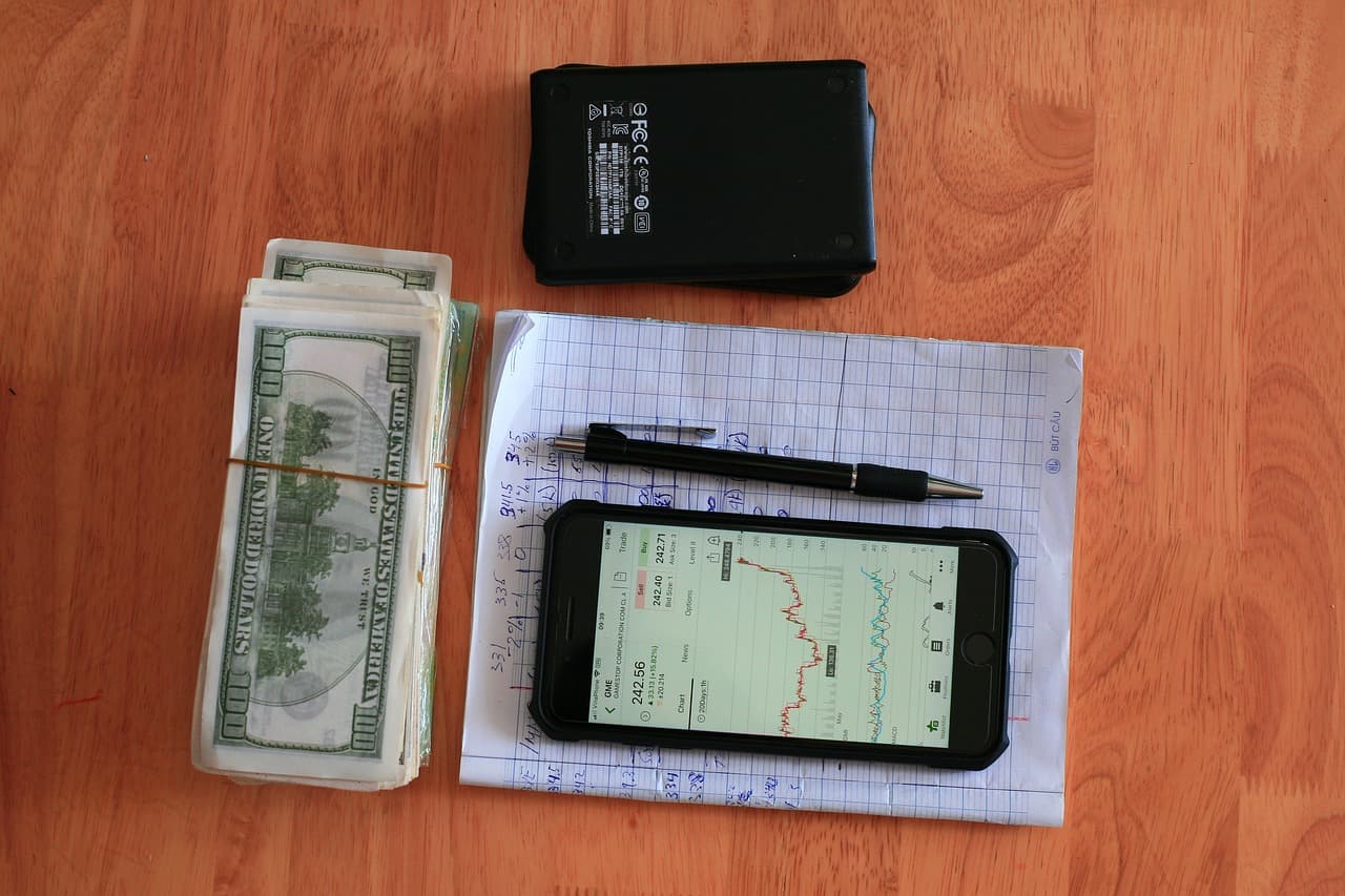 A piece of paper with writing on it that has a pen and an iphone showing a candle chart of the crypto markets resting on top.