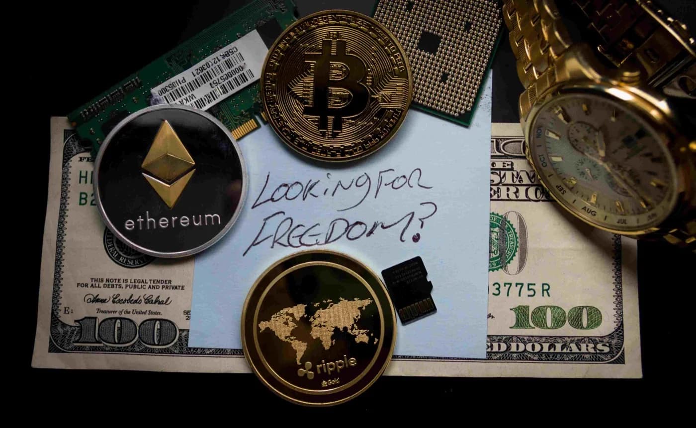 Three cryptocurrency coins resting on top of a one hundred dollar bill with a piece of paper on top and a gold watch next to it.