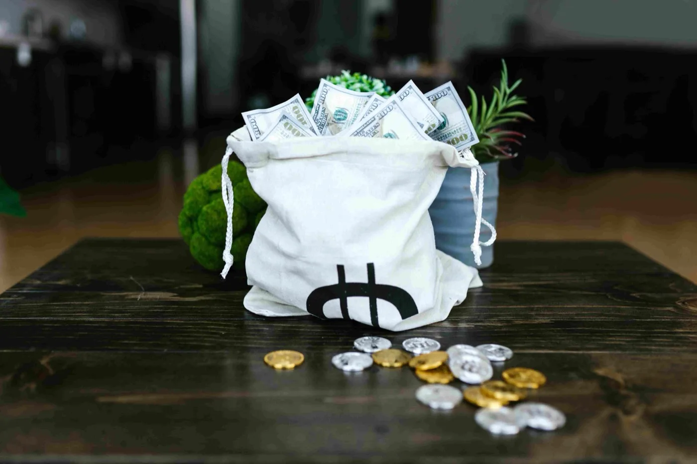 A white money bag filled with stacks of cash next to some gold and silver coin showing the Bitcoin logo all on a brown table.