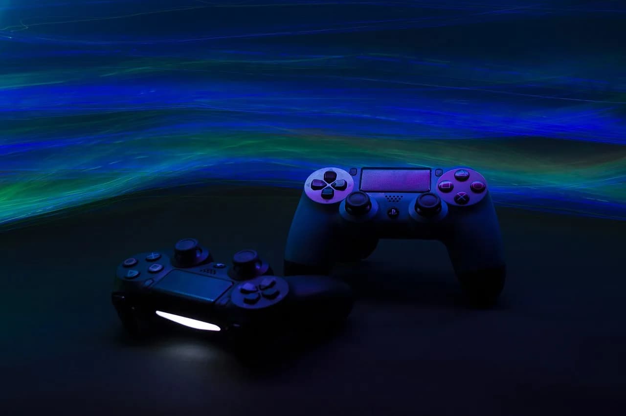 Two video game controllers on a black table in a dark room with a green and blue flow of lines that are lit up passing by them.