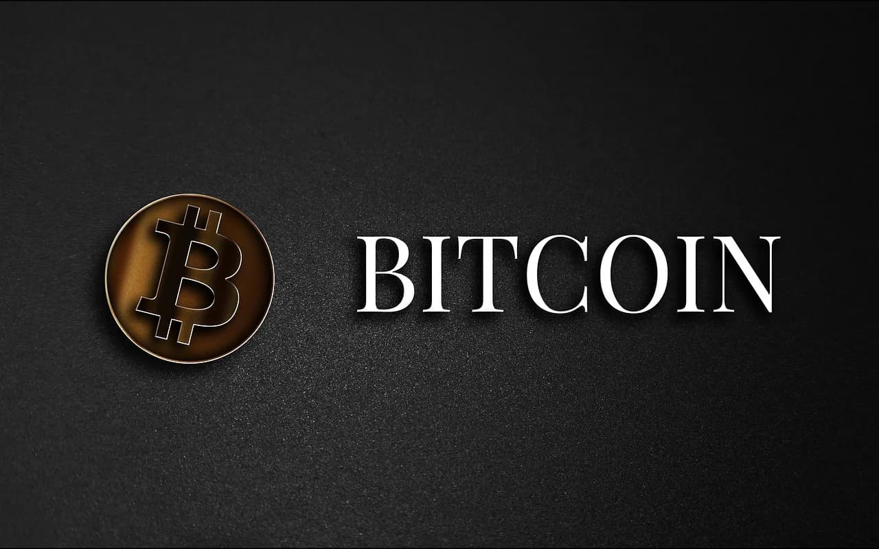 A golden coin with the Bitcoin logo on it in black next to the word Bitcoin all in front of a shadowy dark gray background.