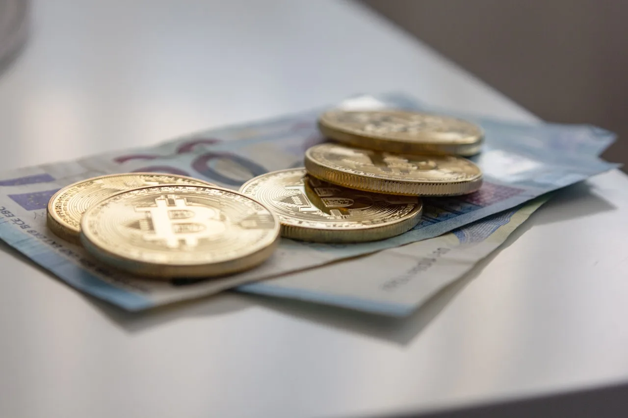 A stack of green currency bills on a white table with multiple golden coins resting on top all with the Bitcoin logo showing.