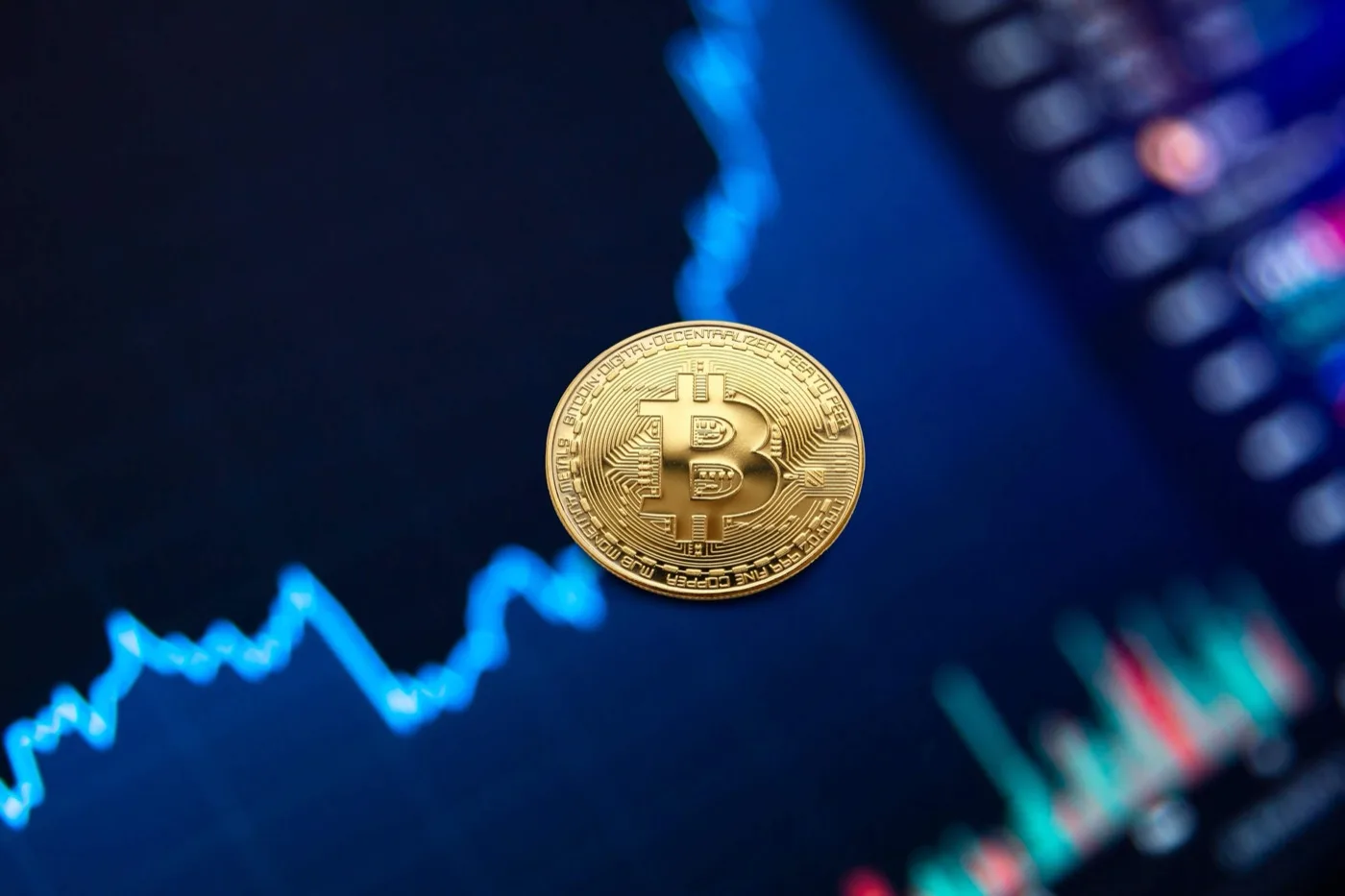 Bitcoin sitting in front of a computer screen that has the crypto market graph that is in the color blue.