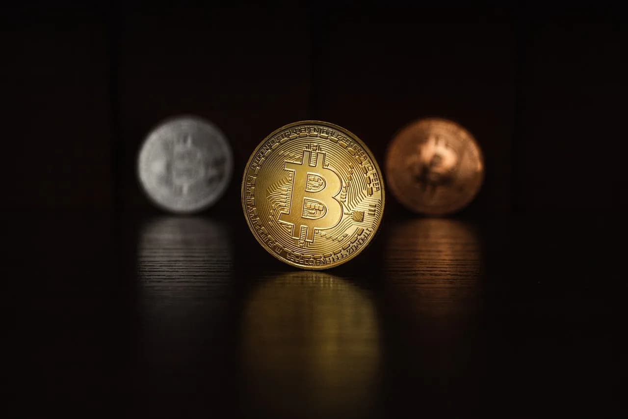 A golden coin with the bitcoin logo on it in front of a silver and bronze coins with the logo as well in a darkly lit room.