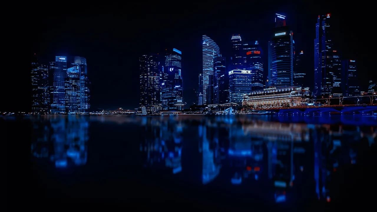 A city on the water is lit up with blue lights and each building has its own white and yellow room lights on in random windows.