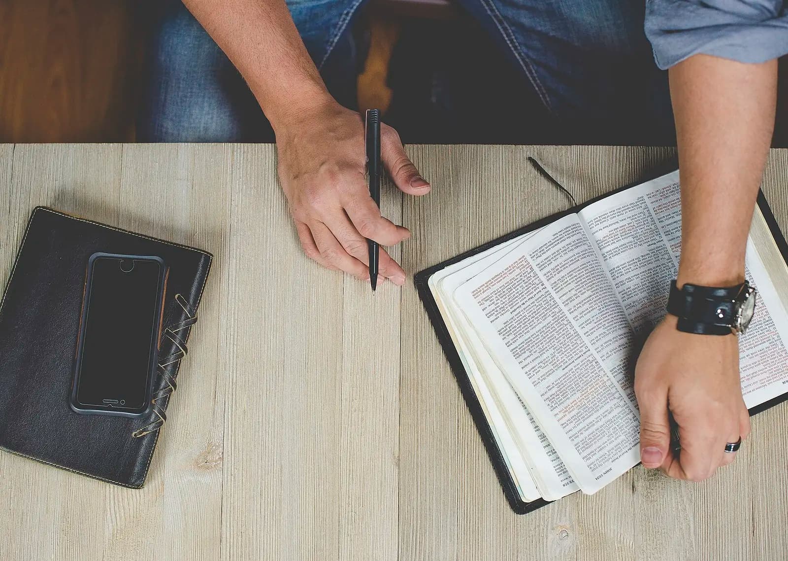Hands resting beside an open Bible and a phone on a wooden table, a quiet Bible app searches moment of study